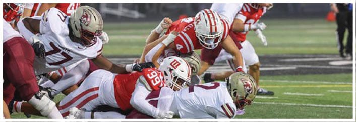 Several football players in red and white uniforms are engaged in a tackle on a field during a game. Description generated by AI