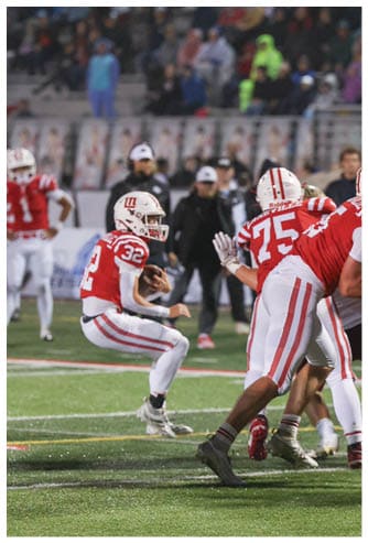 A person wearing a red and white football uniform with the number 32 is holding a football and appears to be preparing to throw it on a grassy field during a game. Description generated by AI