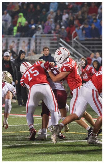Players in red and white uniforms are engaged in a football game on a field with spectators in the background. Description generated by AI