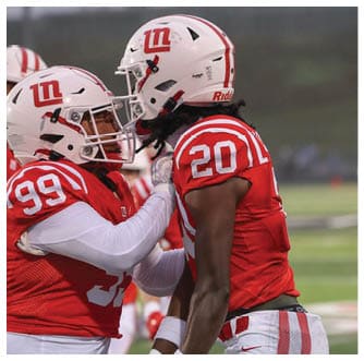 Two people in football uniforms are facing each other on a field, with one wearing a red jersey numbered 20 and a white helmet. Description generated by AI