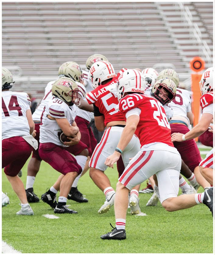 People wearing football uniforms are engaged in a play on a grassy field with empty bleachers in the background. Description generated by AI