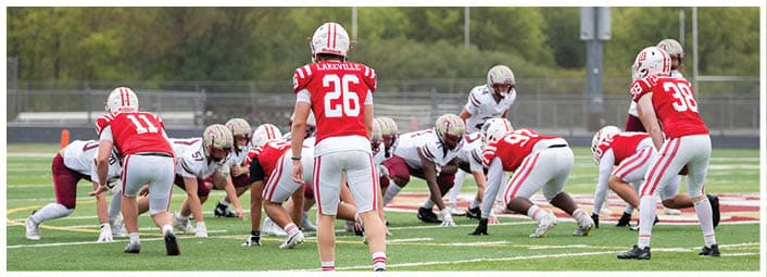 People in red and white football uniforms are lined up on a field facing opponents in white and maroon uniforms, preparing for a play. Description generated by AI