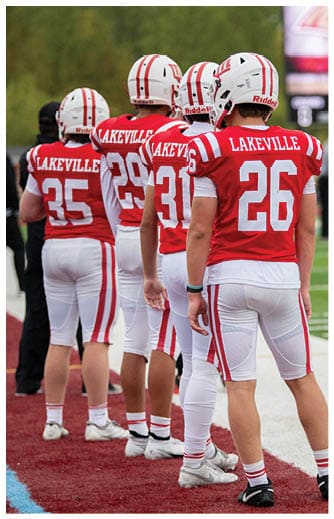 Several people wearing red and white football uniforms with the word Lakeville on the back are standing on a sports field. Description generated by AI
