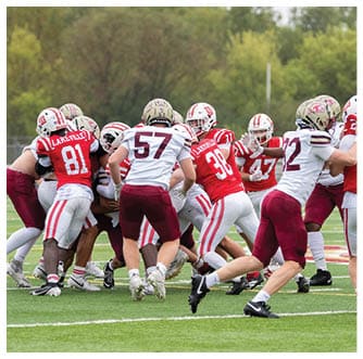 Several people in football uniforms are engaged in a close play on a grassy field with some players wearing red and white and others in white and maroon. Description generated by AI