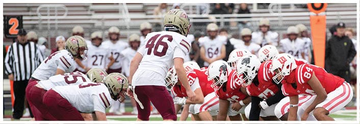 Two opposing football teams in red and white uniforms are lined up facing each other on a field, ready for a play, with referees and people in the background. Description generated by AI