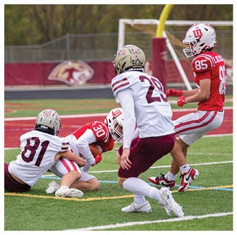 Two people in red and white football uniforms are on the ground while two people in white and maroon uniforms approach them on a football field near the end zone. Description generated by AI