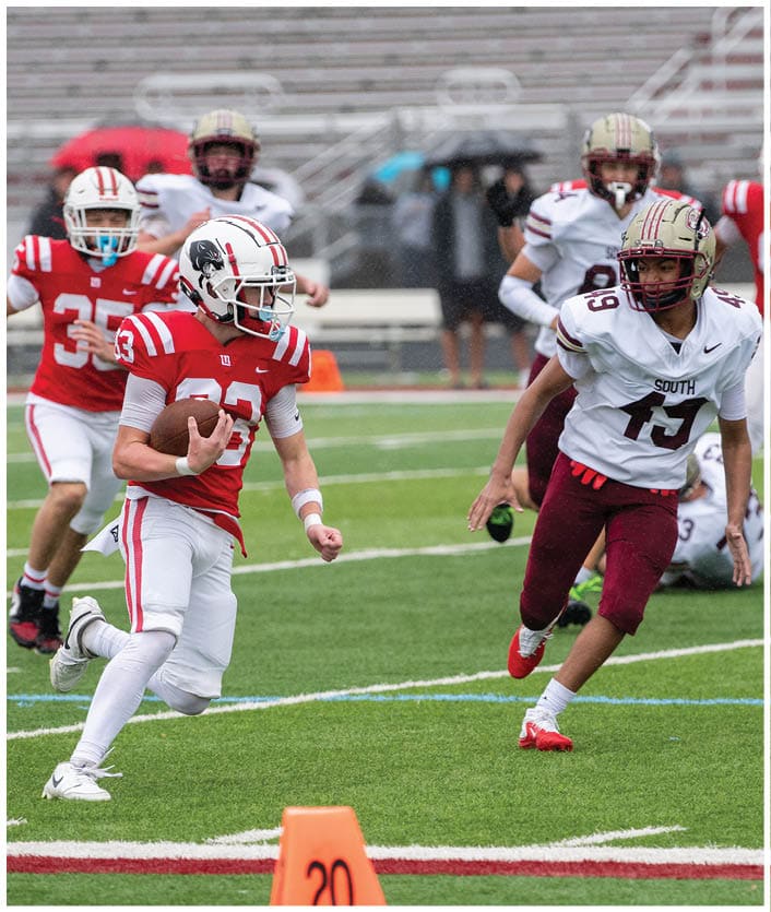 A person in a red and white football uniform is running with the ball on a green field while other players in red and white and maroon and white uniforms chase behind. Description generated by AI