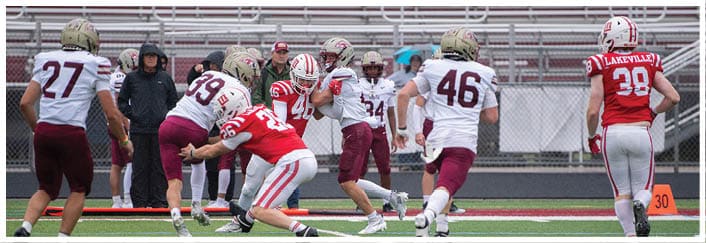 Players in red and white and maroon and white uniforms are actively engaged in a football game on a green field with empty bleachers in the background. Description generated by AI
