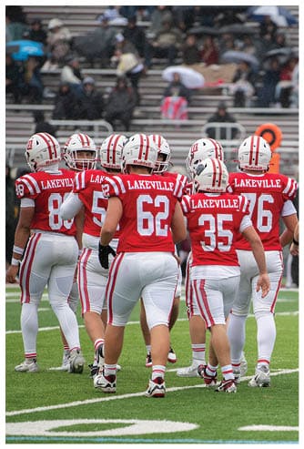A group of people wearing red and white football uniforms with “Lakeville" on the back are standing together on a green football field with bleachers and spectators in the background. Description generated by AI