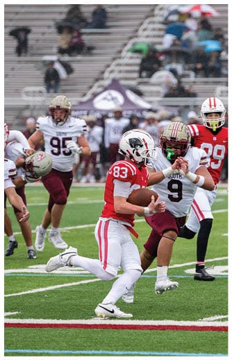 A person in a red and white football uniform is running with the ball while being pursued by two people in white and maroon football uniforms on a green field. Description generated by AI