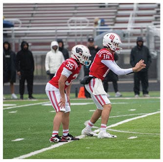 Two people in red and white football uniforms and helmets are positioned on a field, with one person appearing to prepare to snap the ball and the other standing nearby. Description generated by AI