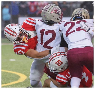 Two people in football uniforms are engaged in a tackle during a game, with one wearing a white jersey numbered 12 and the other in a red jersey. Description generated by AI