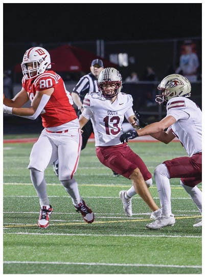 Two people in football uniforms are engaged in a play on a field with one wearing a white and maroon uniform and the other in a red and white uniform, while a referee watches in the background. Description generated by AI