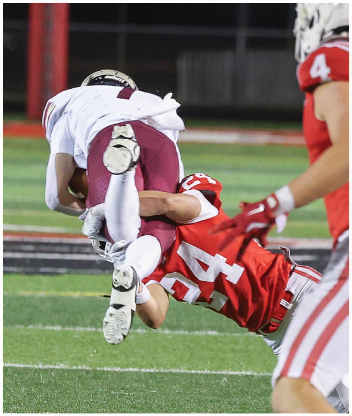 A person in a red football uniform is tackling another person in a white and maroon football uniform during a game on a field. Description generated by AI