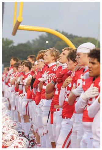 A line of people wearing red and white football uniforms stand side by side with their hands over their hearts near a football goalpost. Description generated by AI