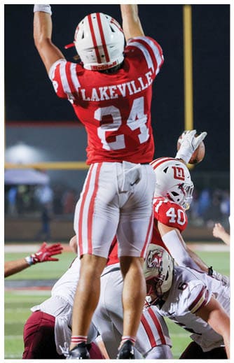 A person wearing a red and white football uniform with the number 24 and the word Lakeville is standing on a football field while other players are engaged in a play near the ground. Description generated by AI