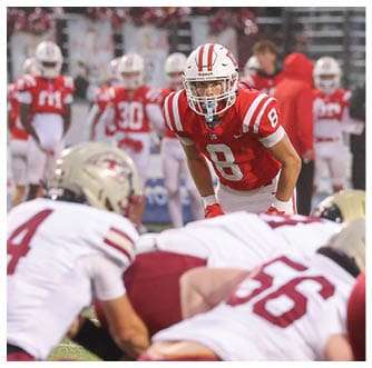 A person in a red football uniform with the number 8 is crouched and focused on players in white and maroon uniforms preparing for a play on a football field. Description generated by AI