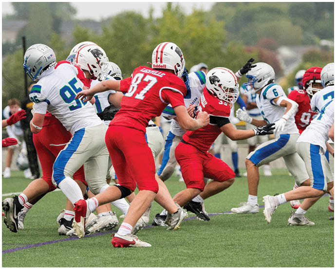People wearing red and white football uniforms are actively playing on a green field with one person in white holding a football and others in red attempting to block or tackle. Description generated by AI