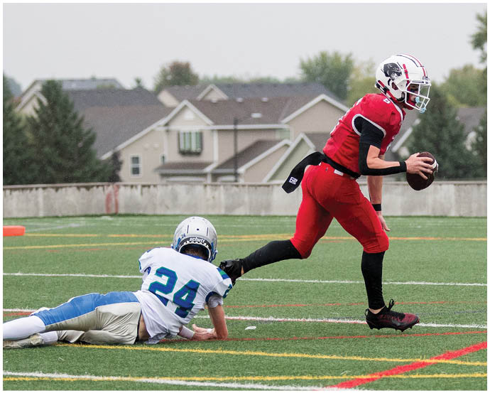 A person in red sports gear is running with a football while another person in white and blue sports gear is on the ground reaching out to try to stop them on a grassy field with houses and trees in the background. Description generated by AI