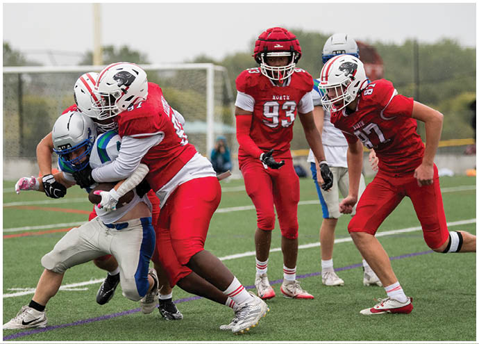 Four people in football uniforms are on a field with two players in red tackling a player in white and blue while another player in red watches nearby. Description generated by AI