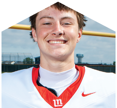 A person wearing a white and red sports jersey is smiling outdoors with a football goalpost and a field in the background. Description generated by AI