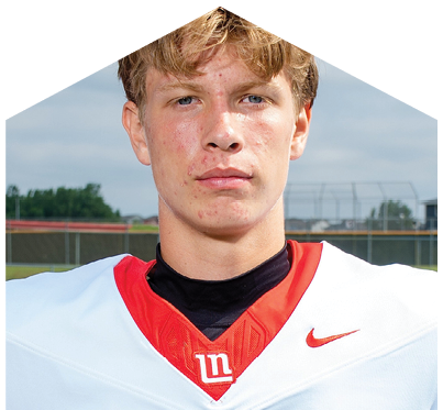 A person with short wavy hair wearing a white and red football jersey stands outdoors on a sports field with a fence and buildings in the background. Description generated by AI