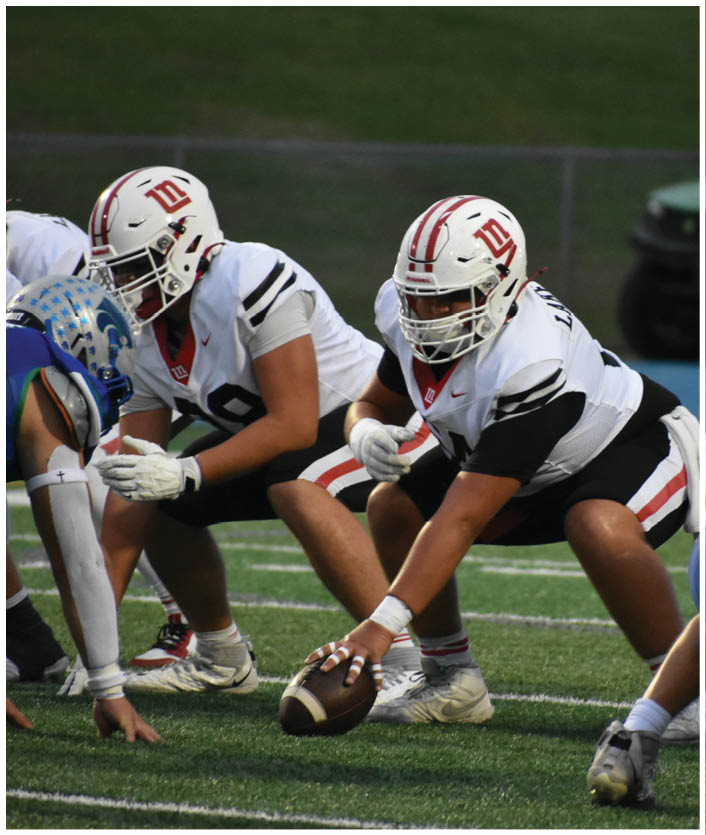 Two people in football uniforms are positioned on a field with one holding a football ready to snap it. Description generated by AI