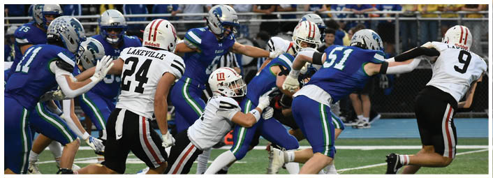 People wearing blue and white football uniforms are actively engaged in a play on a green field with a blue track in the background. Description generated by AI