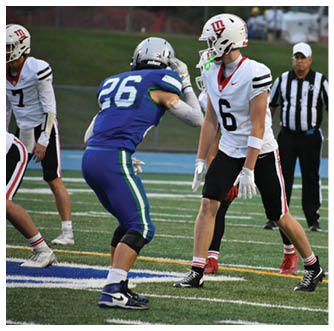 Two people in football uniforms are facing each other on a field with one person in a blue uniform and the other in a white uniform, while a referee stands in the background. Description generated by AI