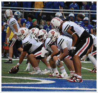A group of football players in white uniforms with helmets are lined up on a field preparing for a play while players in blue uniforms stand in the background. Description generated by AI