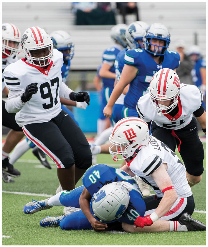 Several people in football uniforms are engaged in a tackle on a green field with empty bleachers in the background. Description generated by AI