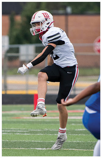 A person in a white and black football uniform is kicking a football on a green field with another player partially visible nearby. Description generated by AI