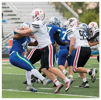 Players in white and blue football uniforms are engaged in a tackle on a green field with empty bleachers in the background. Description generated by AI
