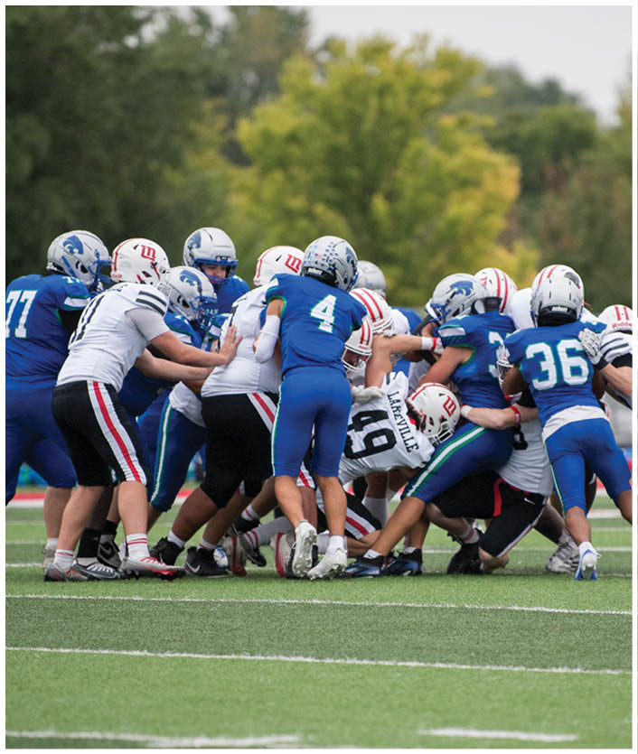 People wearing football helmets and uniforms are engaged in a close, intense play on a grassy field with trees in the background. Description generated by AI
