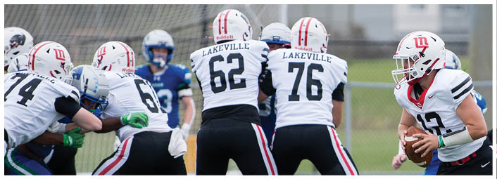 Three people in white football uniforms with “Lakeville" on the back are on a field, with one person holding a football and others positioned nearby. Description generated by AI
