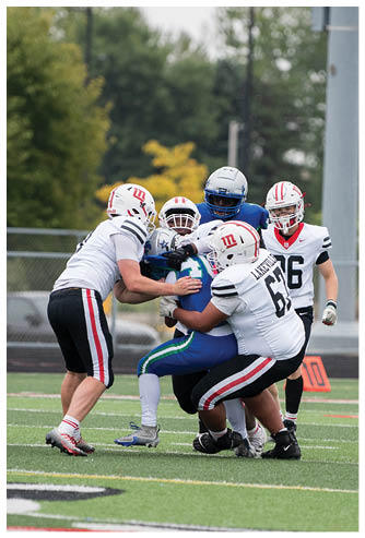Four people in football uniforms are engaged in a tackle on a grassy field with a blurred background of trees and a fence. Description generated by AI