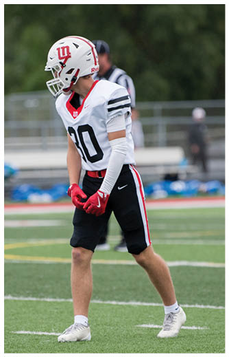 A person wearing a sports uniform and white cleats is running on a green football field near the sideline with blurred spectators and cars in the background. Description generated by AI