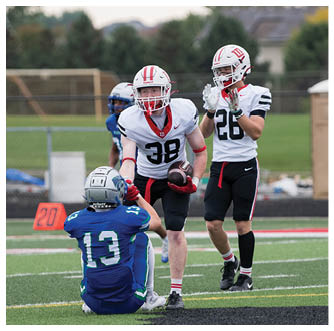Two people in white football uniforms stand on a field with one holding a football while a person in a blue uniform sits on the ground facing them. Description generated by AI