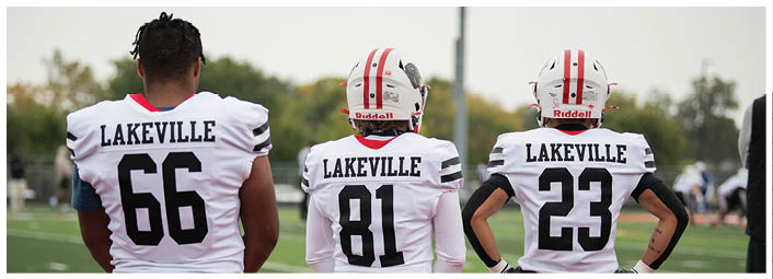 Three people wearing white football jerseys with the name Lakeville and different numbers stand on a field facing away from the camera. Description generated by AI