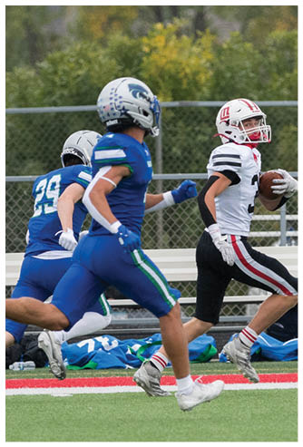 A person in a white and black football uniform is running with the ball while two people in blue football uniforms chase them on a field with a chain-link fence and greenery in the background. Description generated by AI