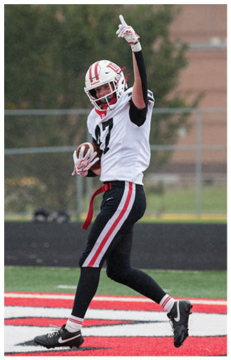 A person in a white football uniform with black and red stripes is jumping on a red and white field while raising one arm with a pointed finger. Description generated by AI