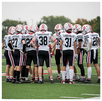 A group of football players wearing white helmets and white jerseys with “Lakeville" on the back are huddled together on a green field. Description generated by AI