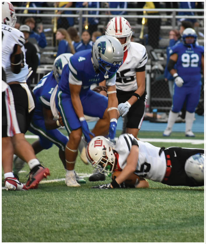 People in football uniforms are engaged in a play on a field with one person in white lying on the ground while others in blue and white stand nearby. Description generated by AI