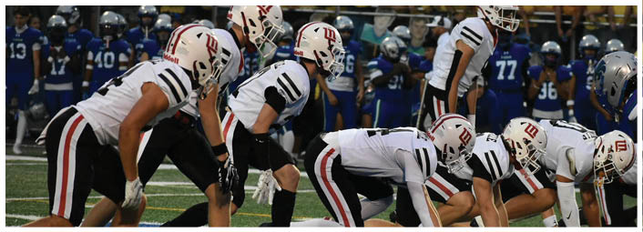 A group of people in white sports uniforms and helmets are lined up in a football formation on a field with opponents in blue uniforms visible in the background. Description generated by AI