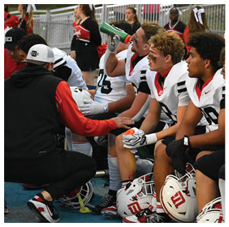 People in football uniforms sit on a bench while one person in a black and red jacket kneels in front of them, and one player drinks from a green water bottle. Description generated by AI