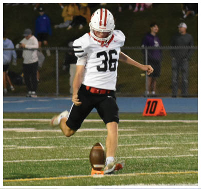 A person wearing a white football uniform is kicking a football on a field while another person in a similar uniform runs nearby with spectators watching in the background. Description generated by AI