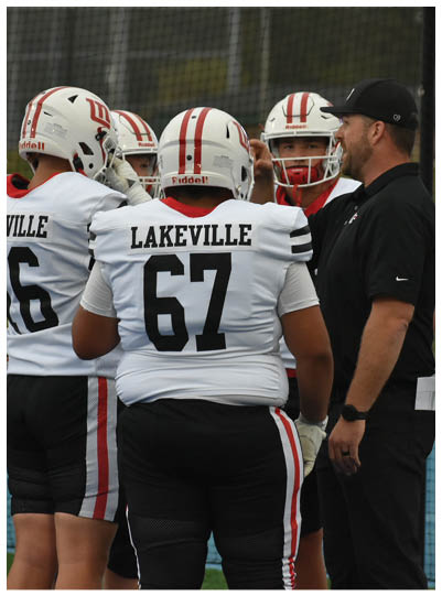 A group of people wearing white football uniforms with “Lakeville" on the back are gathered around a person in black athletic clothing and a black cap on a sports field. Description generated by AI