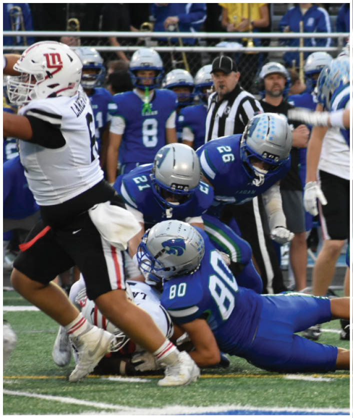 Several football players in blue uniforms are tackling a player in a white uniform on a field with a blue and white logo. Description generated by AI
