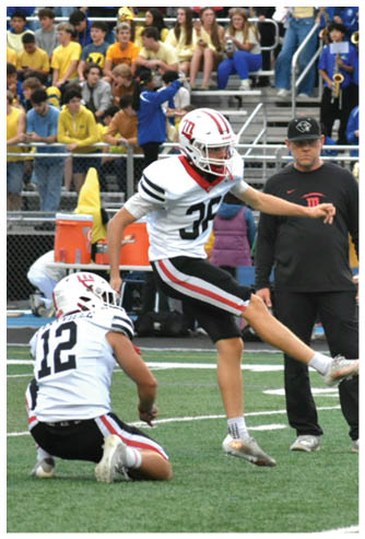 A person in a white football uniform with the number 36 is kicking a football on a field while a person in black clothing stands nearby and a crowd watches from the bleachers. Description generated by AI