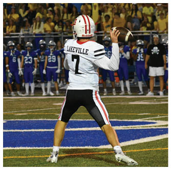A person wearing a white football uniform with the name Lakeville and number 7 is preparing to throw a football on a field with players and spectators in the background. Description generated by AI
